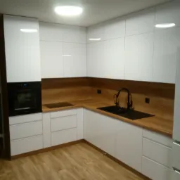 Nowoczesna kuchnia w kształcie litery L z białymi, lakierowanymi frontami, drewnianym blatem i czarną armaturą, oświetlona okrągłą lampą sufitową.