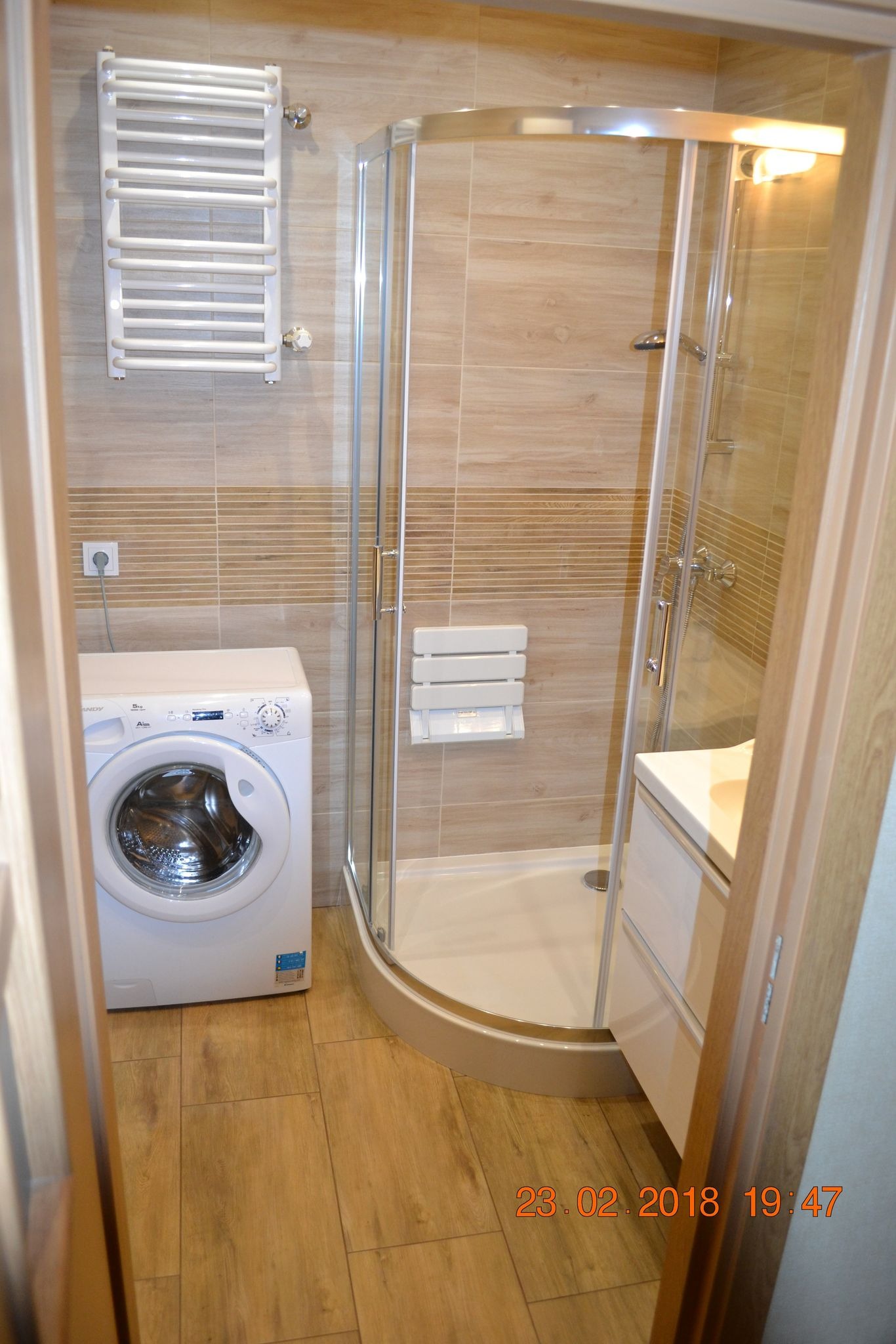 Ukończona łazienka z kabiną prysznicową narożną, umywalką z szafką, pralką i grzejnikiem drabinkowym, wykończona płytkami w odcieniach beżu i brązu, z drewnopodobną podłogą.