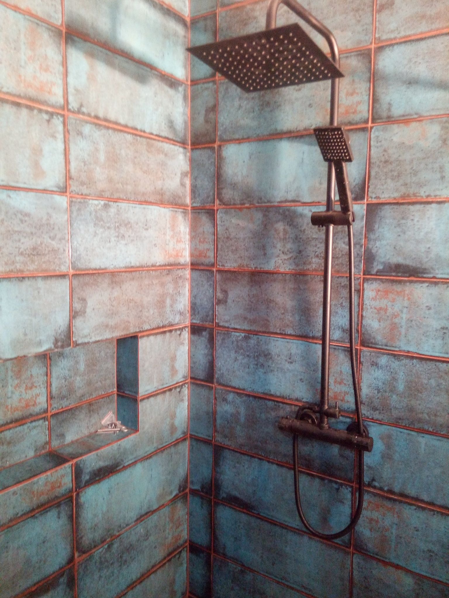 Kabina prysznicowa wyłożona turkusowymi, prostokątnymi płytkami z rdzawymi fugami, wyposażona w czarną deszczownicę i baterię z termostatem.