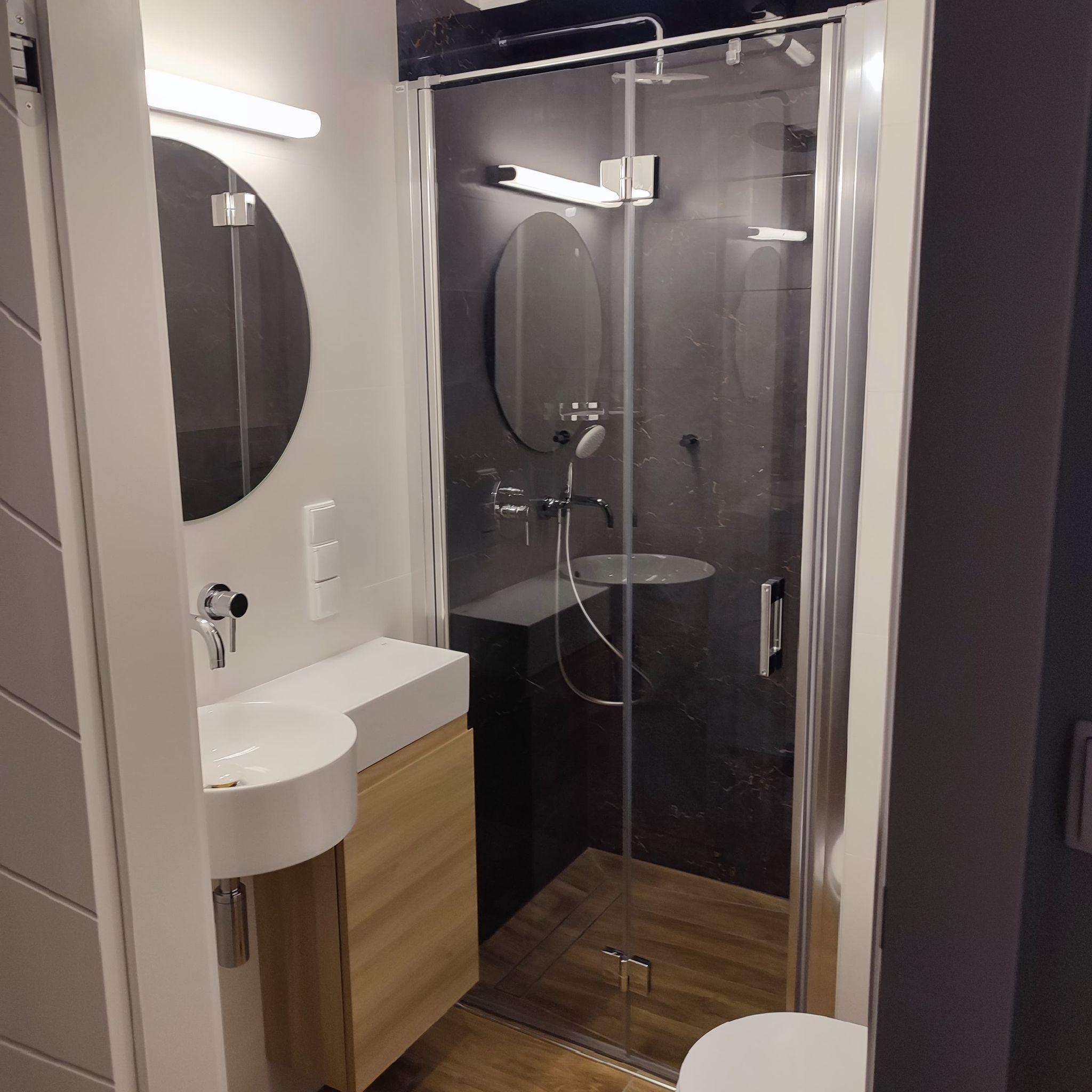 Nowoczesna łazienka z kabiną prysznicową typu walk-in z czarnymi płytkami imitującymi marmur, okrągłym lustrem z podświetleniem LED i umywalką nablatową na szafce z drewnopodobnym frontem.