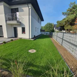 Nowoczesny dom jednorodzinny z zadbanym trawnikiem z rolki, obok ogrodzenie panelowe z podmurówką, widok z perspektywy trawnika w kierunku domu.