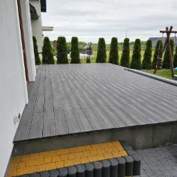 Szary taras kompozytowy z widoczną strukturą drewna, obrzeżem z żółtej kostki i rzędem tuj w tle, przylegający do białego budynku.