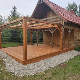 SPARKTECH Apostolos Aleksandros Dodis - Drewniany taras z pergolą przy domku, otoczony kamieniami i zielenią. Widoczne detale konstrukcji, oświetlenie naturalne, neutralna kolorystyka drewna.