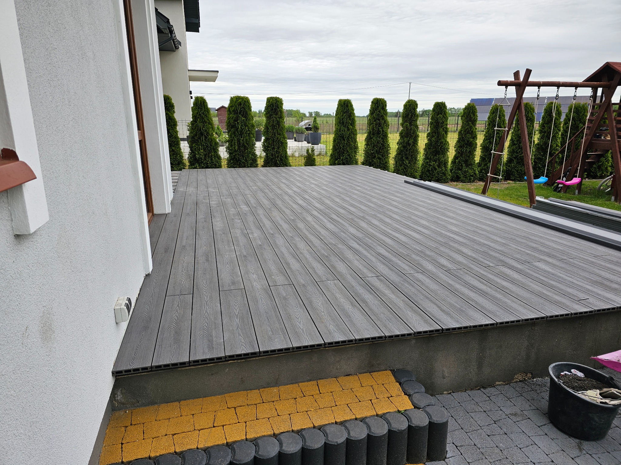 Szary taras kompozytowy z widoczną strukturą drewna, obrzeżem z żółtej kostki i rzędem tuj w tle, przylegający do białego budynku.