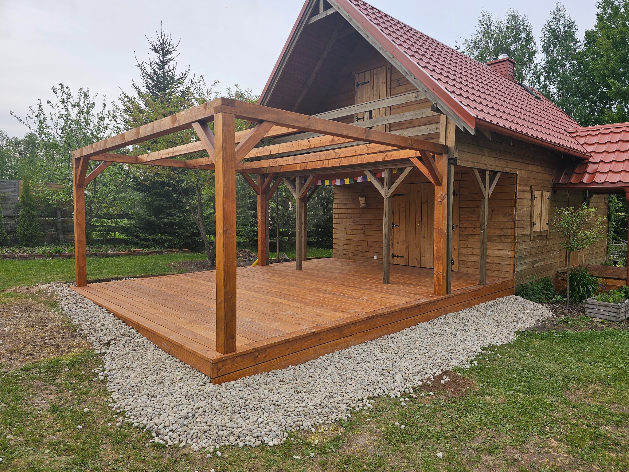 Drewniany taras z pergolą przy domku, otoczony kamieniami i zielenią. Widoczne detale konstrukcji, oświetlenie naturalne, neutralna kolorystyka drewna.