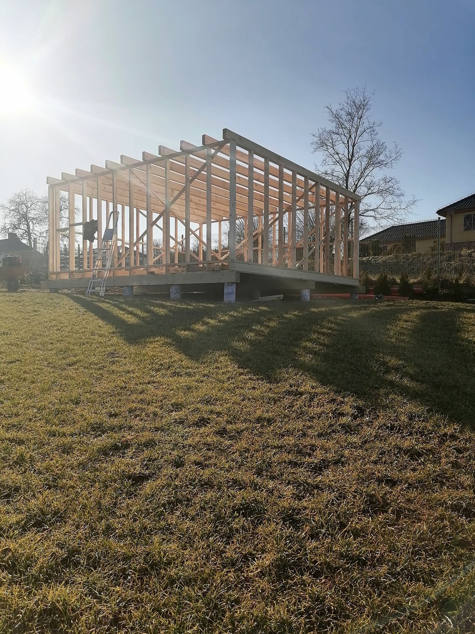 Wznoszenie konstrukcji szkieletowej budynku na trawniku w słoneczny dzień. Widoczny drewniany szkielet, drabina i częściowo dom w tle. Wczesny etap budowy.