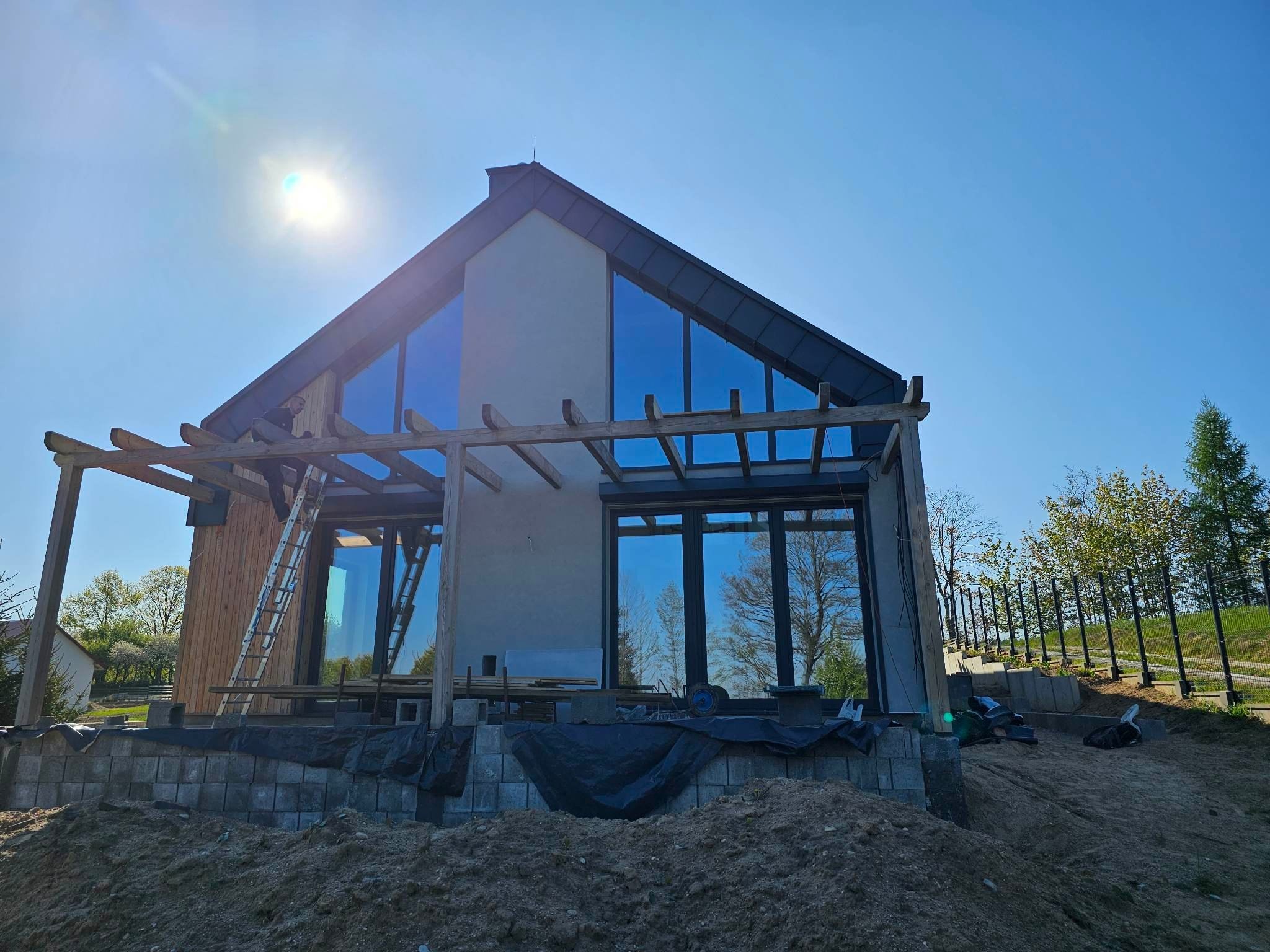 Nowoczesny dom szkieletowy z dużymi oknami i drewnianą pergolą w trakcie budowy. Widoczne elementy konstrukcyjne i rusztowanie. Słoneczny dzień, błękitne niebo.