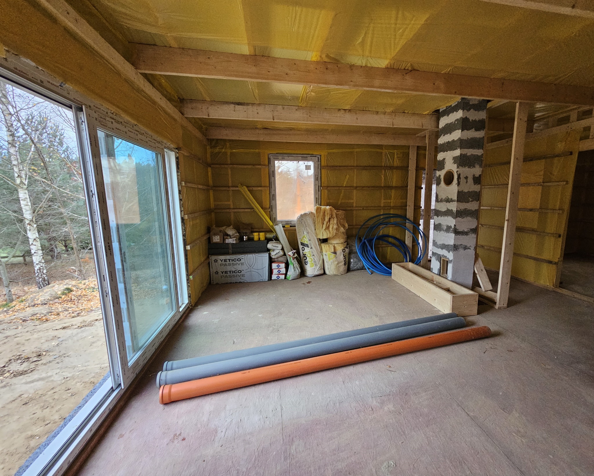 Wnętrze domu szkieletowego w trakcie budowy, widoczne ocieplenie ścian wełną mineralną, okno, komin i rury kanalizacyjne. Surowy stan, brak wykończenia.