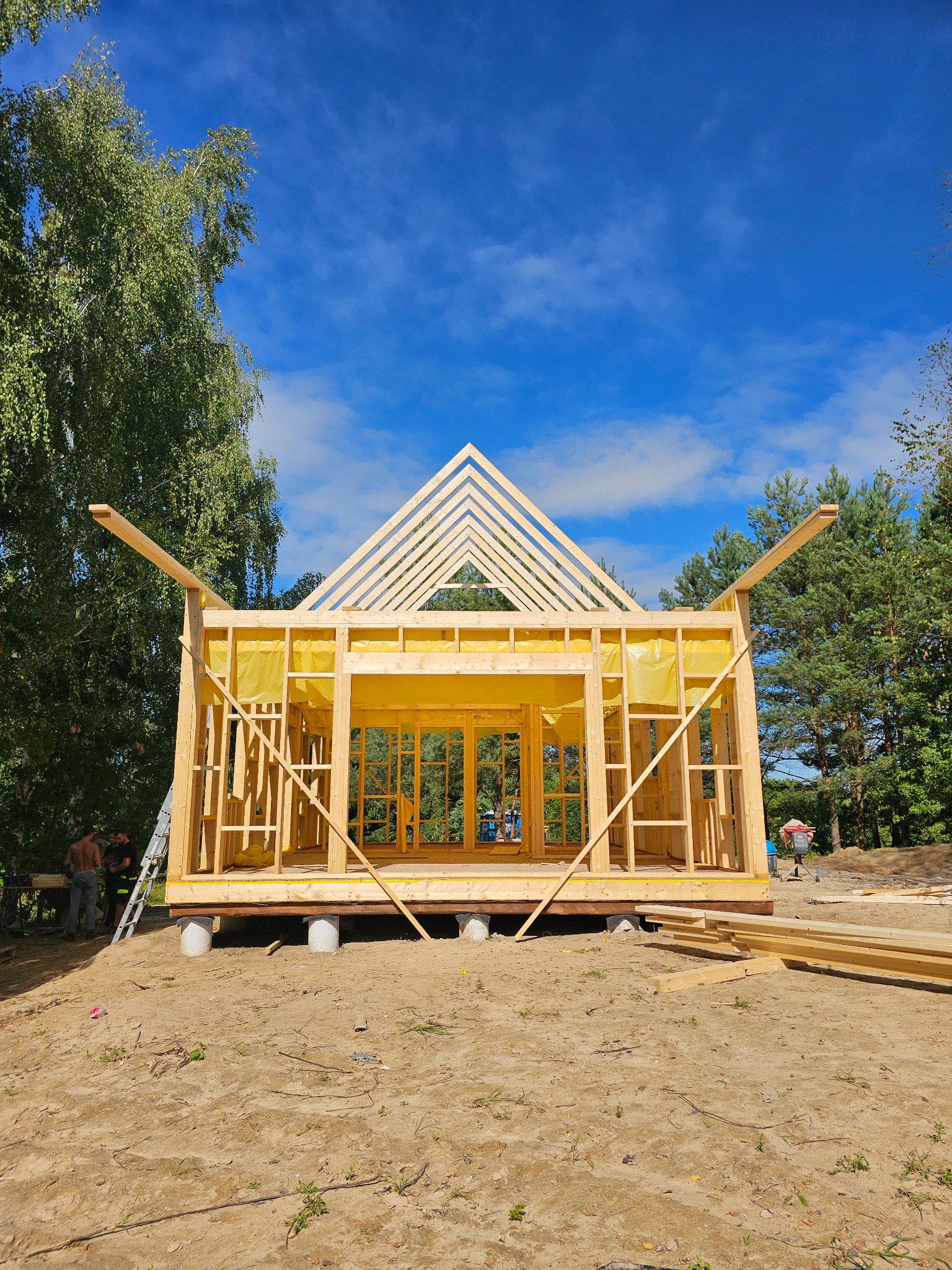 Konstrukcja szkieletowa domu w trakcie budowy, widoczne drewniane elementy i żółta folia izolacyjna, na tle błękitnego nieba i drzew. Częściowo widoczni pracownicy.