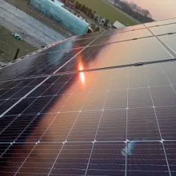 Panele słoneczne na dachu budynku, z odbiciem zachodzącego słońca i widocznym zabrudzeniem na jednym z paneli. W tle zabudowania gospodarcze i pole.