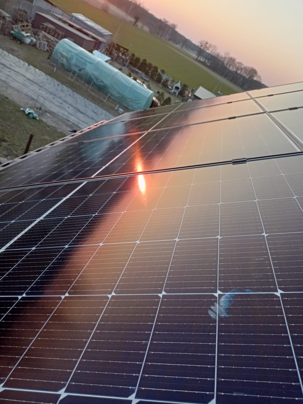 Panele słoneczne na dachu budynku, z odbiciem zachodzącego słońca i widocznym zabrudzeniem na jednym z paneli. W tle zabudowania gospodarcze i pole.