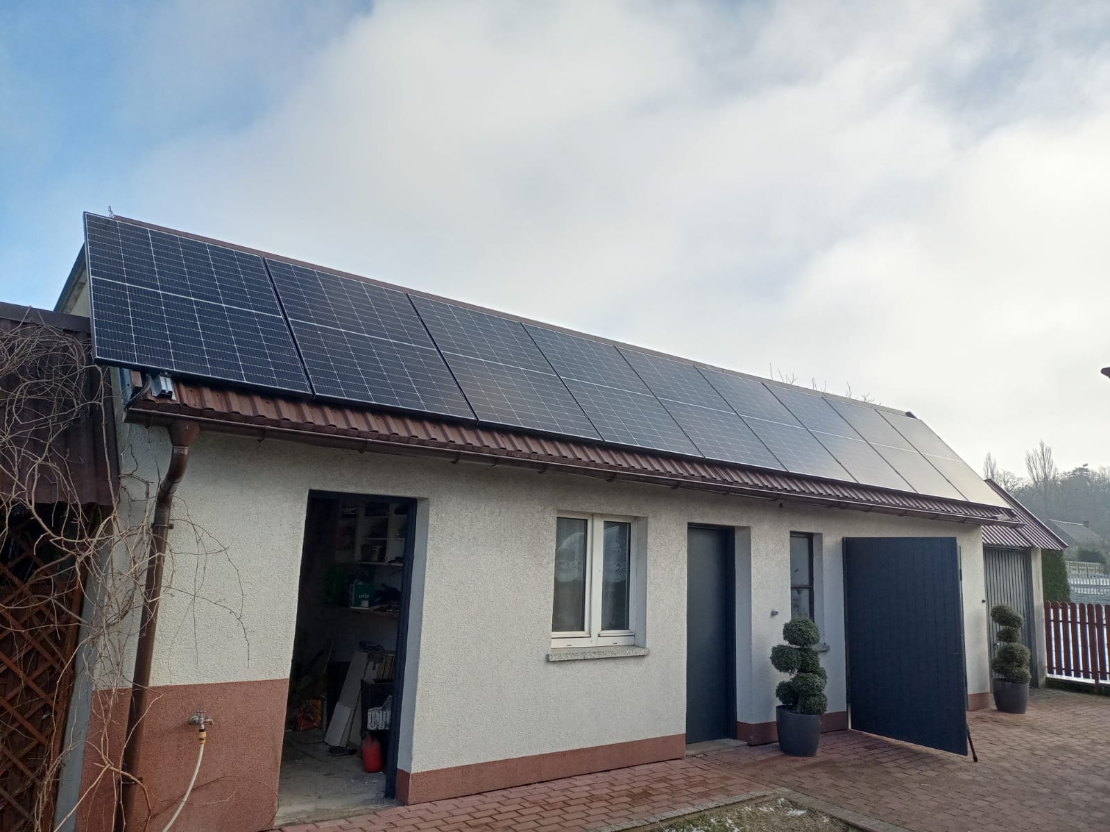 Panele słoneczne na dachu budynku gospodarczego z brązową dachówką, widoczne rynny i fragment posesji z kostki brukowej