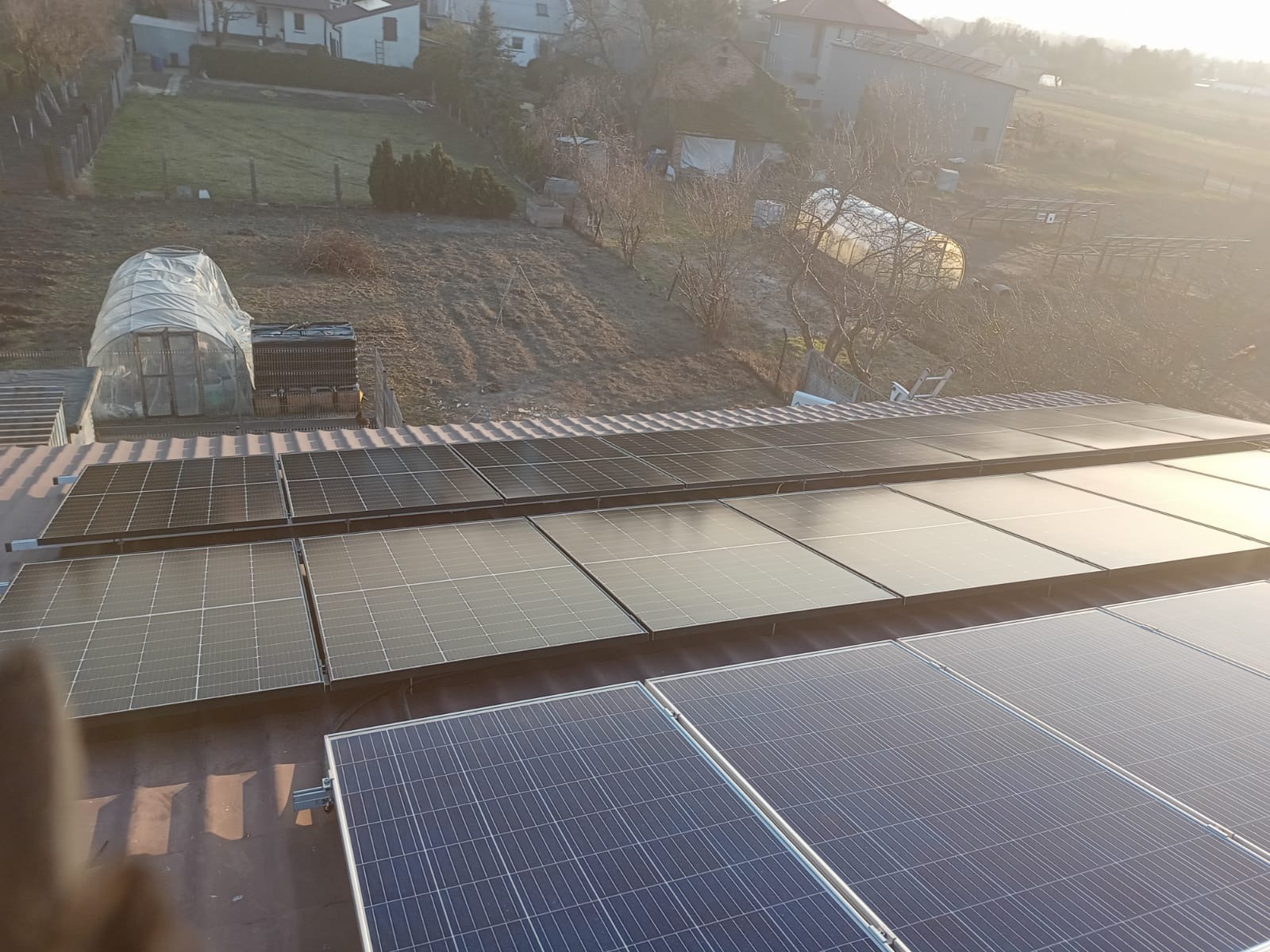 Instalacja fotowoltaiczna na dachu domu jednorodzinnego, widok z góry, w tle szklarnia i zabudowa mieszkalna w promieniach wschodzącego słońca.