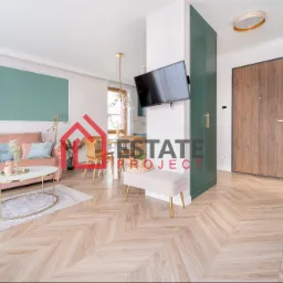 Jasny salon z parkietem w jodełkę, różową sofą, okrągłym stolikiem kawowym i zieloną zabudową meblową przy wejściu.