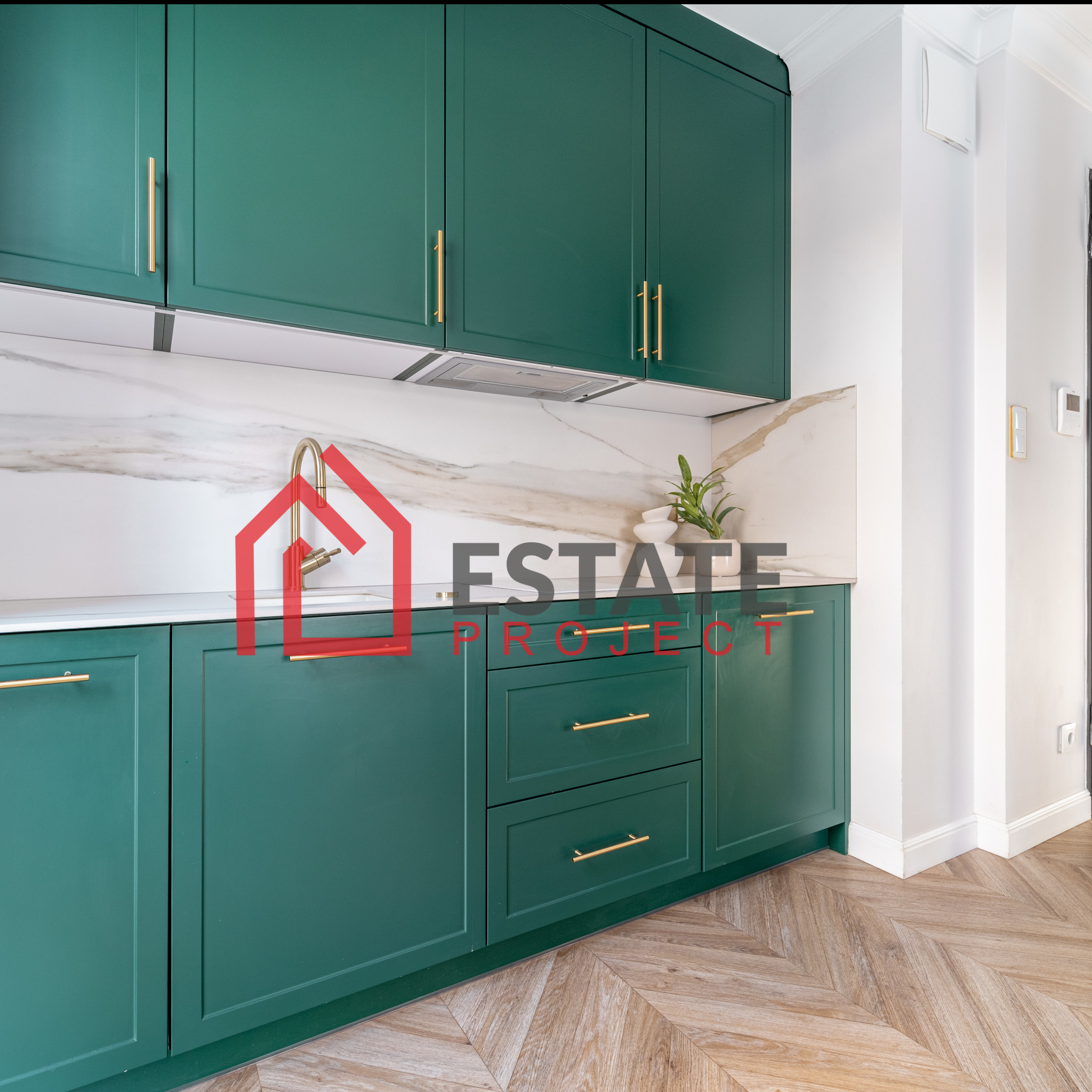 Szmaragdowa zabudowa kuchenna z białym blatem i marmurowym backsplashiem, złote uchwyty, parkiet w jodełkę.