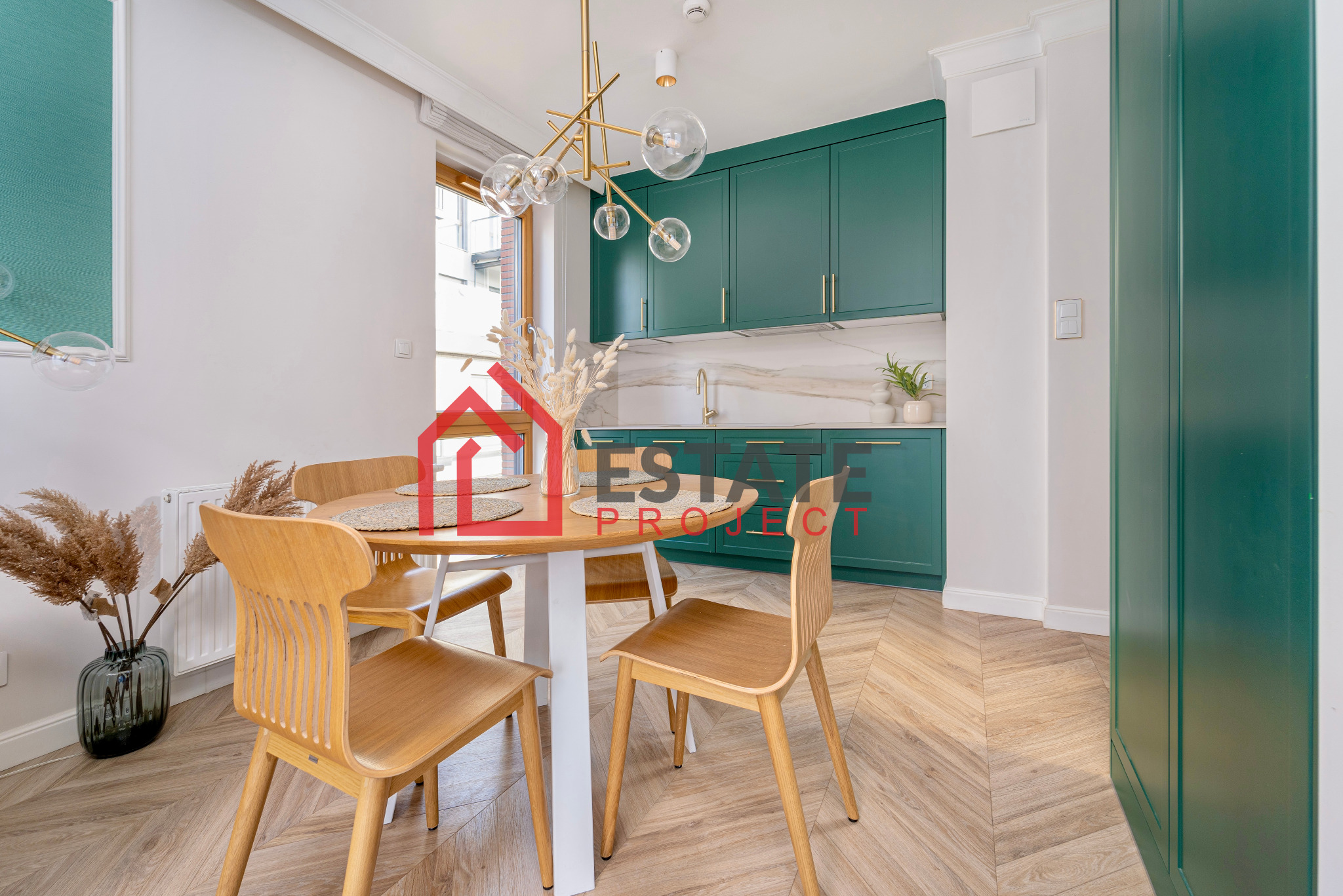 Jasne drewniane krzesła przy okrągłym stole z suszonymi trawami w wazonie, na tle zielonej zabudowy kuchennej z marmurowym blatem i parkietu ułożonego w jodełkę.