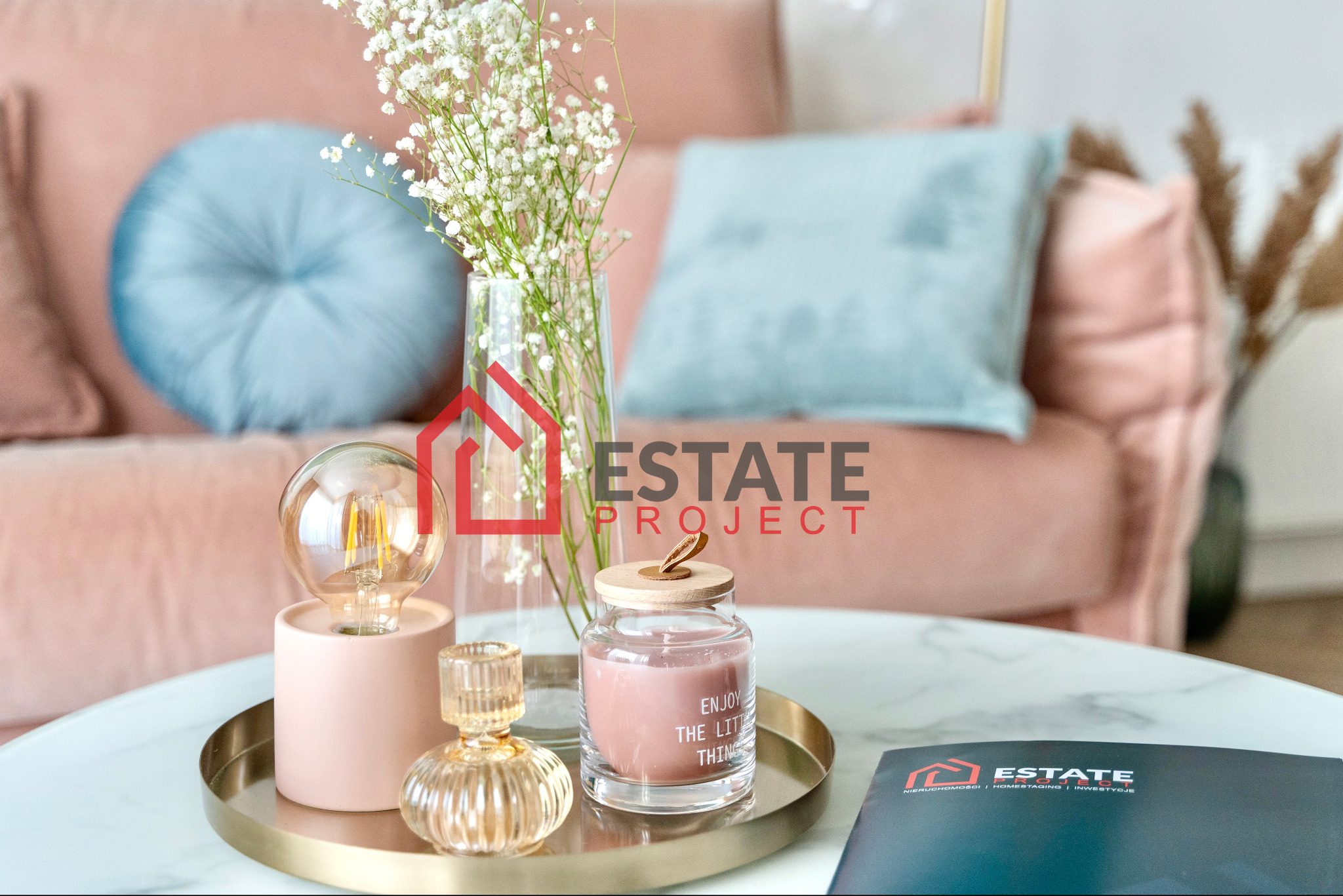 Dekoracyjny stolik kawowy z różowym akcentem: lampka, świeca z napisem 'Enjoy the little things', wazon z gipsówką, broszura firmy Estate Project i pluszowa kanapa w tle.