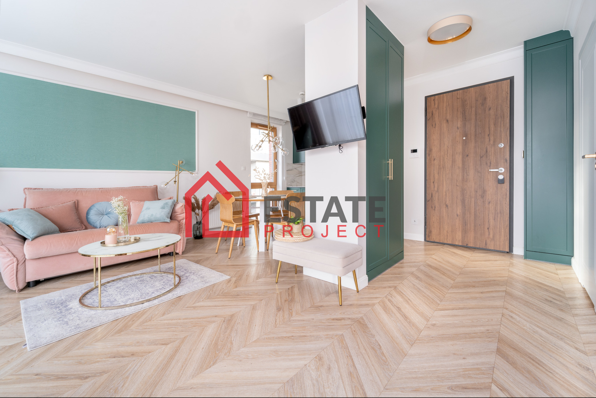 Jasny salon z parkietem w jodełkę, różową sofą, okrągłym stolikiem kawowym i zieloną zabudową meblową przy wejściu.