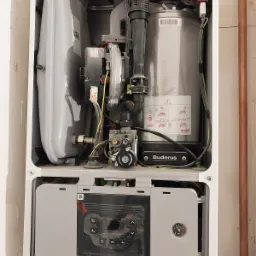 Wnętrze otwartego pieca gazowego marki Buderus z widocznymi elementami instalacji i panelem sterowania, zamontowanego przy ścianie z miedzianą rurą.