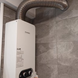 HEAT MASTER SERWIS URZĄDZEŃ GAZOWYCH Marcin Rudzik - Biały piec gazowy Junkers na szarej, marmurkowej ścianie z podłączonym srebrnym, elastycznym przewodem spalinowym, widoczne kosmetyki na półce pod piecem.