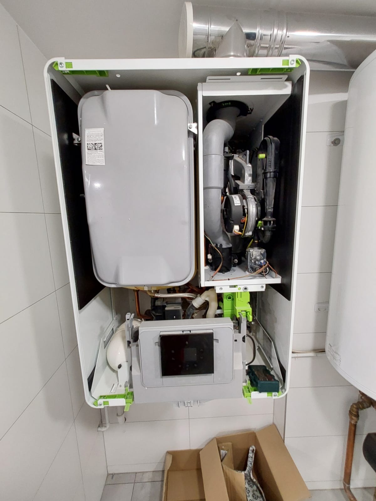 Otwarty piec gazowy z widocznymi elementami wewnętrznymi, zamontowany na ścianie wyłożonej białymi płytkami; widoczna kartonowa skrzynka na podłodze.