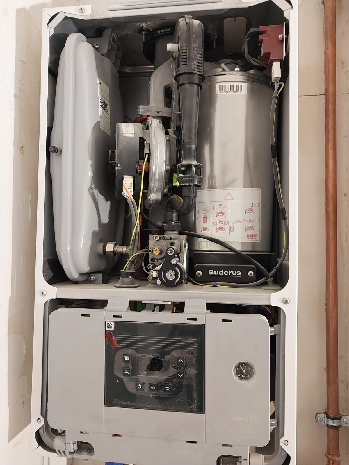 Wnętrze otwartego pieca gazowego marki Buderus z widocznymi elementami instalacji i panelem sterowania, zamontowanego przy ścianie z miedzianą rurą.