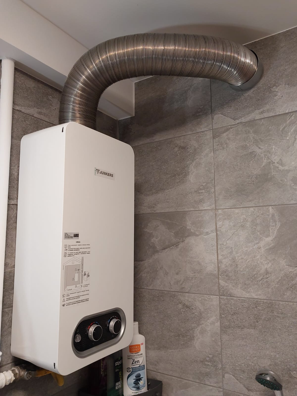 Biały piec gazowy Junkers na szarej, marmurkowej ścianie z podłączonym srebrnym, elastycznym przewodem spalinowym, widoczne kosmetyki na półce pod piecem.