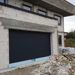 Inter-Decor Aleksander Godziek - Nowoczesna, segmentowa brama garażowa w kolorze grafitowym, zamontowana w surowym stanie budynku z białego bloczka, z widocznym ociepleniem i gruzem budowlanym na podjeździe.