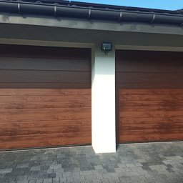 Inter-Decor Aleksander Godziek - Dwie brązowe, segmentowe bramy garażowe z poziomymi panelami imitującymi drewno, oddzielone białym filarem z zamontowanym oświetleniem zewnętrznym, na tle kostki brukowej.