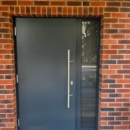 Nowoczesne, grafitowe drzwi wejściowe z długim, srebrnym uchwytem, osadzone w murze z czerwonej cegły z wąskim oknem bocznym.