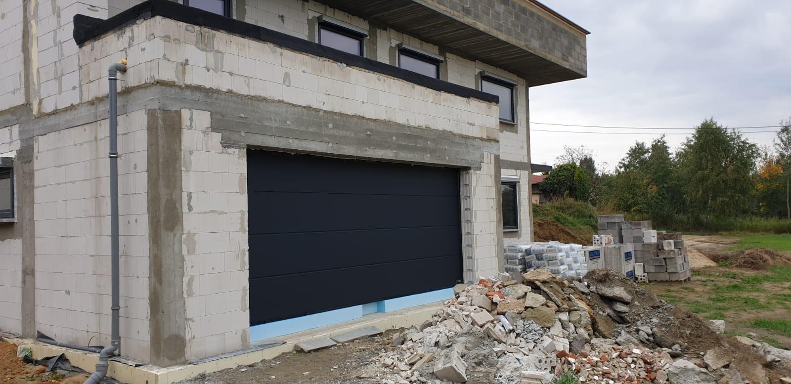 Nowoczesna, segmentowa brama garażowa w kolorze grafitowym, zamontowana w surowym stanie budynku z białego bloczka, z widocznym ociepleniem i gruzem budowlanym na podjeździe.