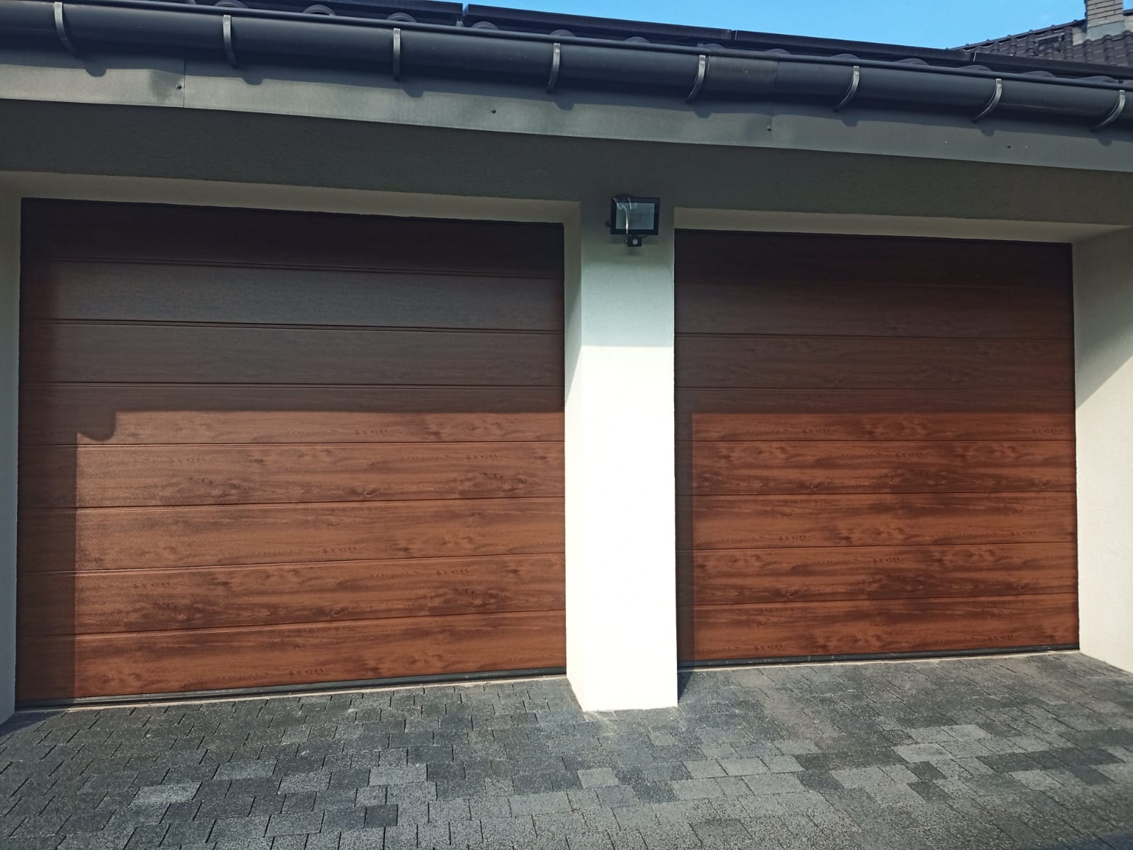 Dwie brązowe, segmentowe bramy garażowe z poziomymi panelami imitującymi drewno, oddzielone białym filarem z zamontowanym oświetleniem zewnętrznym, na tle kostki brukowej.