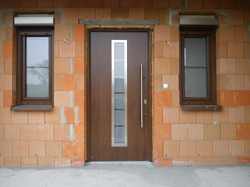 Brązowe drzwi wejściowe z pionowym przeszkleniem i długim uchwytem, zamontowane w surowej ścianie z cegieł kratówek, z oknami po bokach wyposażonymi w zwinięte rolety zewnętrzne.