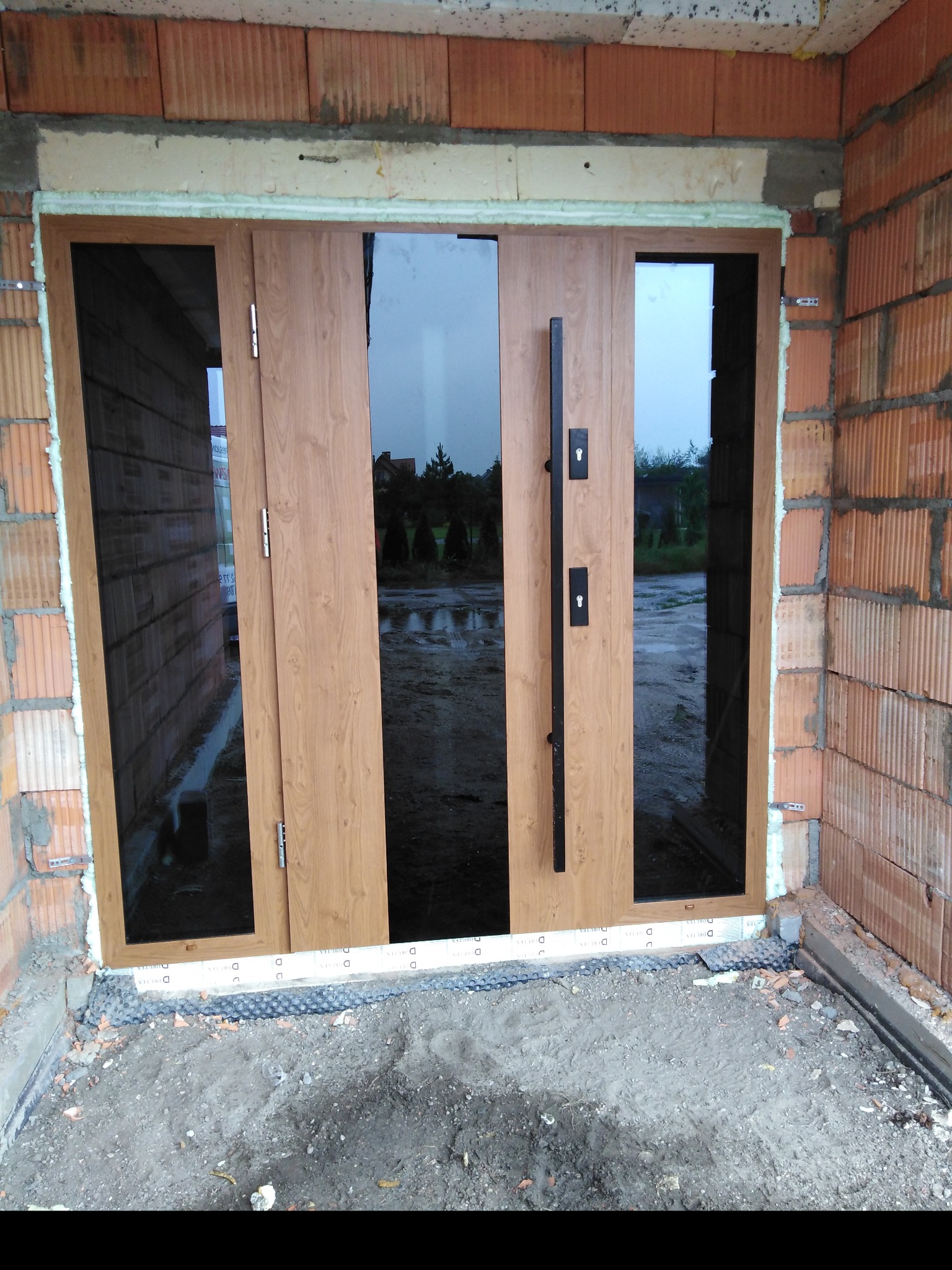 Montaż drzwi zewnętrznych z drewnopodobnym wykończeniem i czarnymi elementami w surowym stanie budynku, widoczne cegły i pianka montażowa.