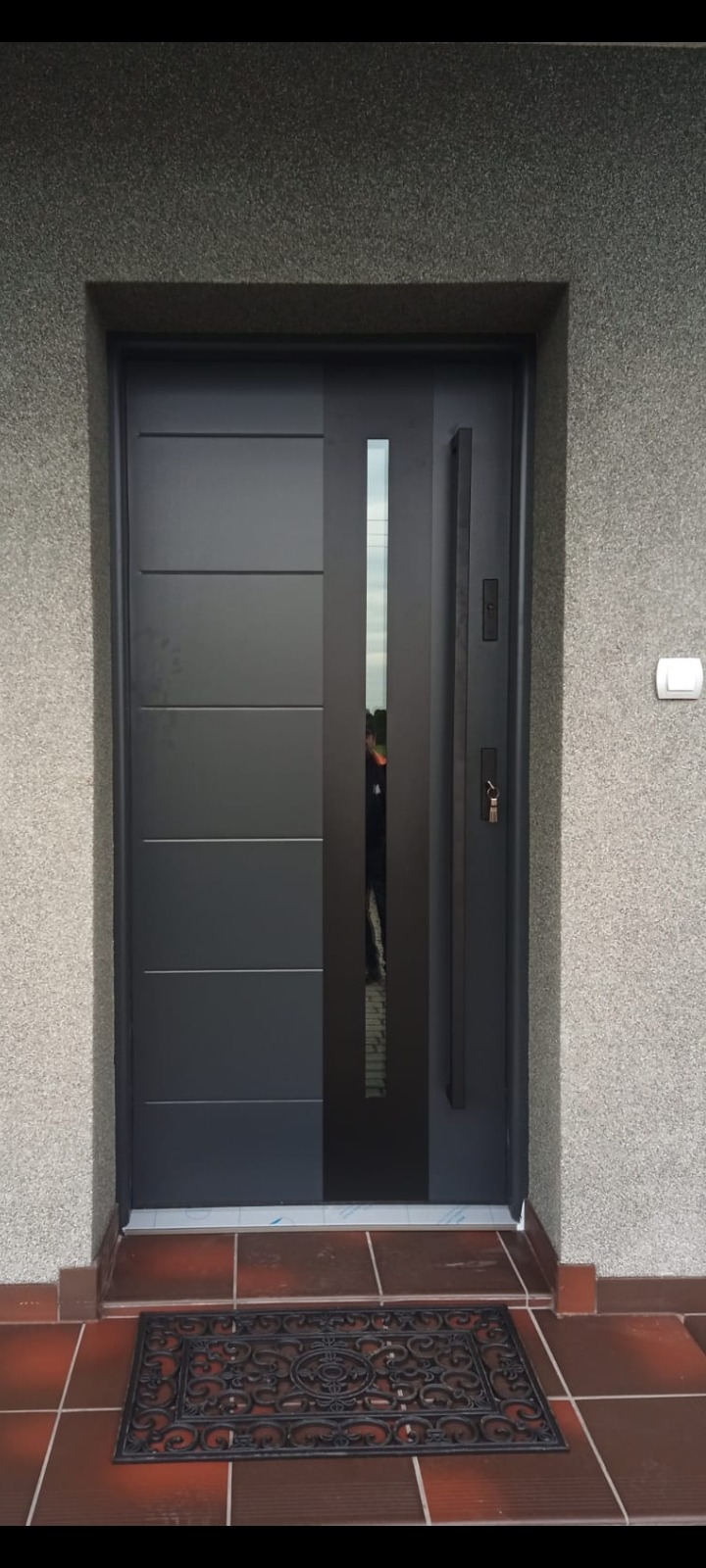 Nowoczesne, grafitowe drzwi wejściowe z pionowym przeszkleniem i długim uchwytem, zamontowane w elewacji o szarej, ziarnistej strukturze; widoczna wycieraczka przed drzwiami.
