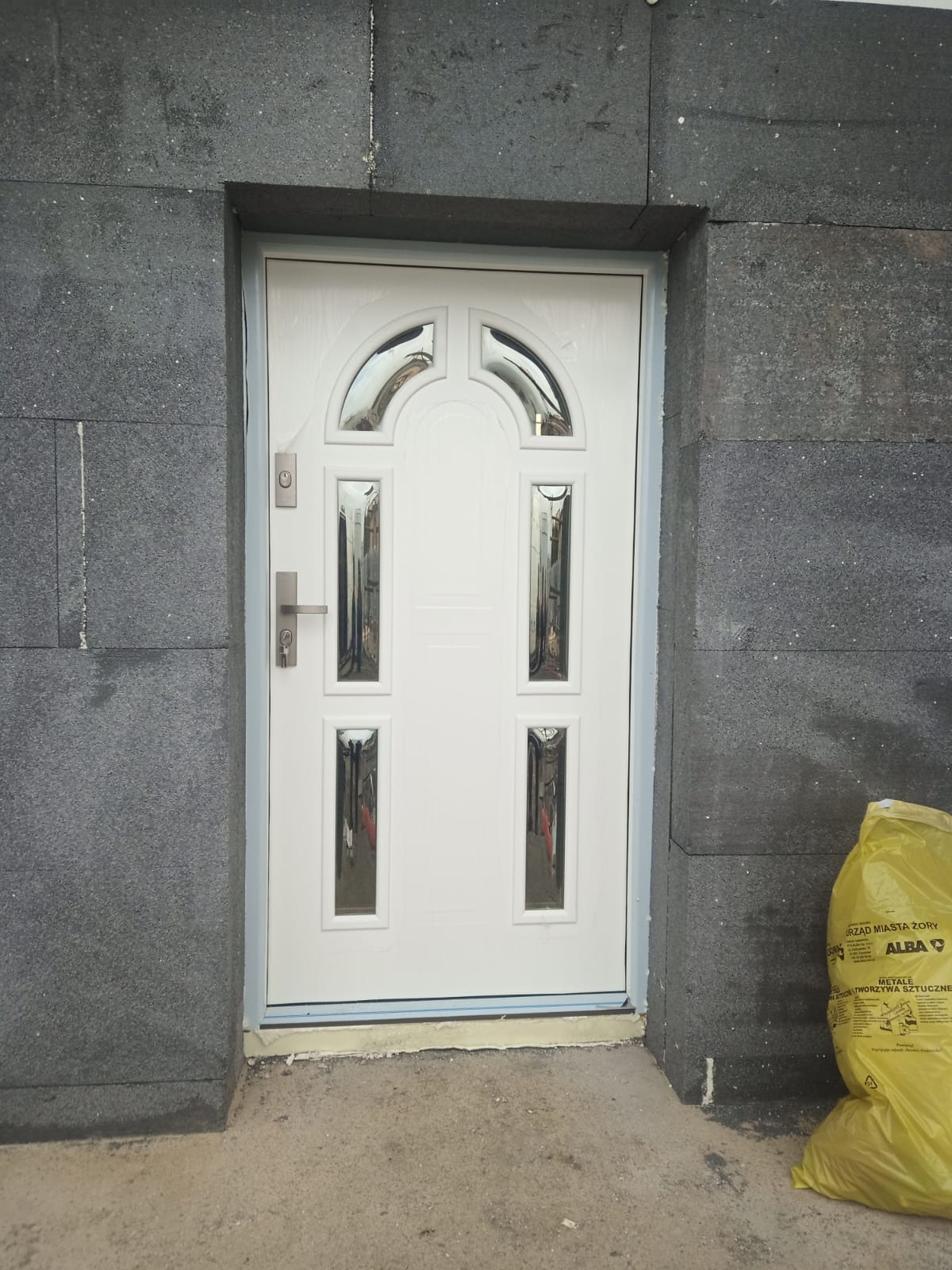 Białe drzwi wejściowe z ozdobnymi szybkami, zamontowane w szarej elewacji, widoczny worek na odpady budowlane.