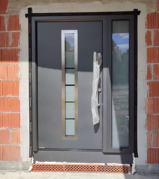 Szare drzwi wejściowe z bocznym oknem, zamontowane w surowej ceglanej ścianie, z widoczną pianką montażową i taśmą uszczelniającą, uchwyt drzwi zabezpieczony siatką ochronną.