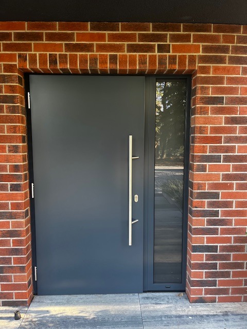 Nowoczesne, grafitowe drzwi wejściowe z długim, srebrnym uchwytem, osadzone w murze z czerwonej cegły z wąskim oknem bocznym.