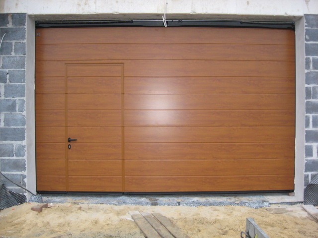 Brązowa segmentowa brama garażowa z dodatkowymi drzwiami wejściowymi, widoczna w trakcie montażu, z surowymi ścianami z bloczków betonowych i piaskiem na posadzce.