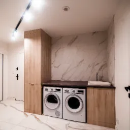 Nowoczesna pralnia w mieszkaniu: pralka i suszarka obok szafki z umywalką, marmurowe płytki na ścianie, oświetlenie szynowe.