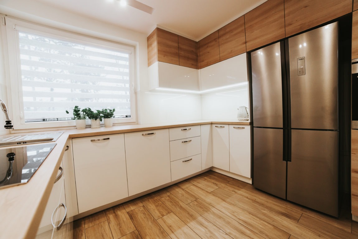 Jasna kuchnia w stylu skandynawskim z białymi frontami szafek, drewnianym blatem i podłogą, oświetleniem LED podszafkowym i dużą, srebrną lodówką side-by-side.