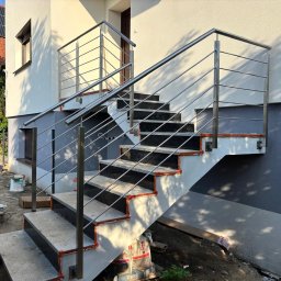 Dagmat - Zewnętrzne schody z czarnymi stopniami i nierdzewną, metalową balustradą, zamontowaną przy jasnym budynku z szarą podmurówką w słoneczny dzień.