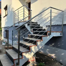 Zewnętrzne schody z czarnymi stopniami i nierdzewną, metalową balustradą, zamontowaną przy jasnym budynku z szarą podmurówką w słoneczny dzień.