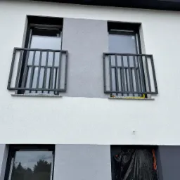 Elewacja budynku z dwoma oknami zabezpieczonymi czarnymi, metalowymi balustradami. Dolne okno zabezpieczone folią. Ściana w odcieniach bieli i szarości.