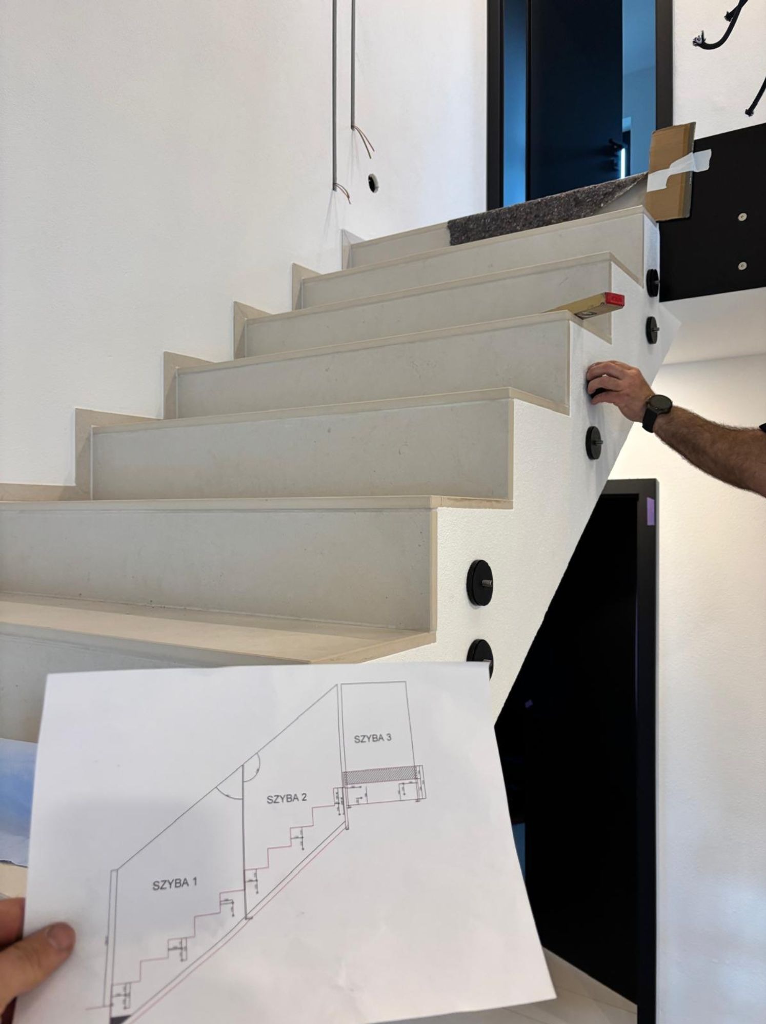 Montaż balustrady szklanej na schodach: widok z dołu na betonowe stopnie z zamontowanymi uchwytami, w tle czarne drzwi i fragment planu montażu szkła trzymanego w dłoni