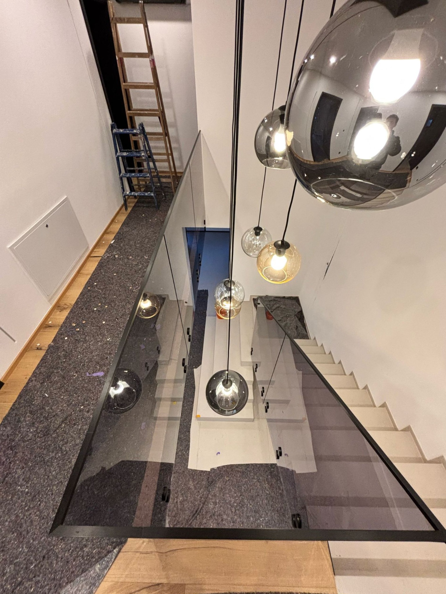 Widok z góry na szklaną balustradę z czarną ramą, zamontowaną na schodach, z widocznymi elementami montażowymi i ochronną szarą matą budowlaną, oraz wiszącymi szklanymi lampami w tle.