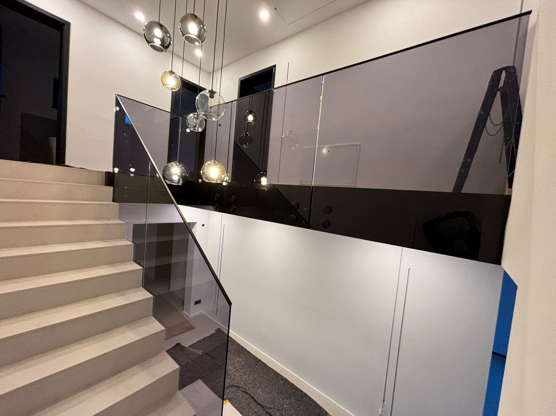 Nowoczesna klatka schodowa z grafitową szklaną balustradą mocowaną na czarnych rotulach, widok z góry na schody i fragment salonu z wiszącymi lampami.