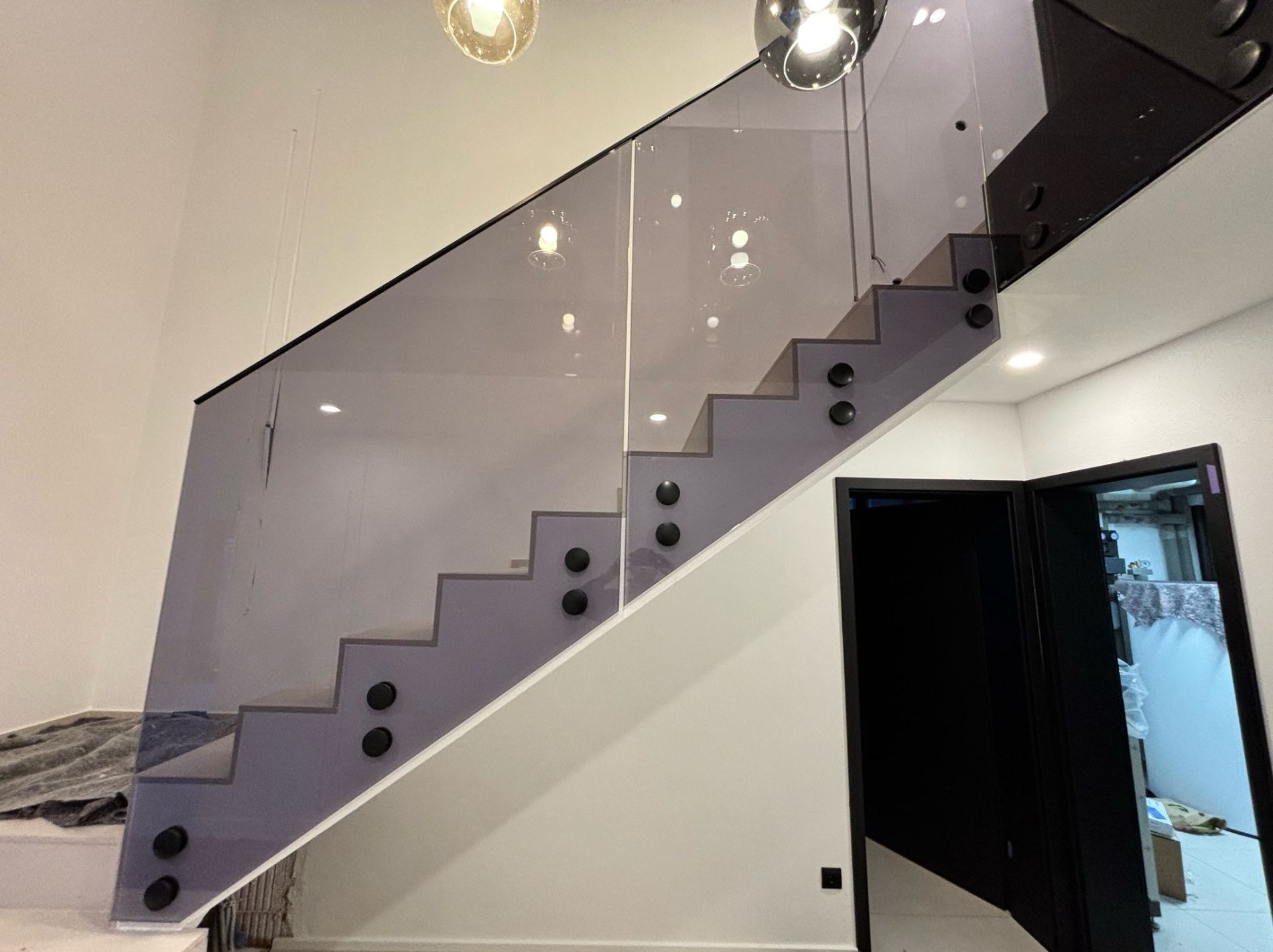 Szklana balustrada schodowa zamocowana na czarnych rotulach, szkło grafitowe, widoczne odbicia lamp sufitowych, schody w kolorze szarym.