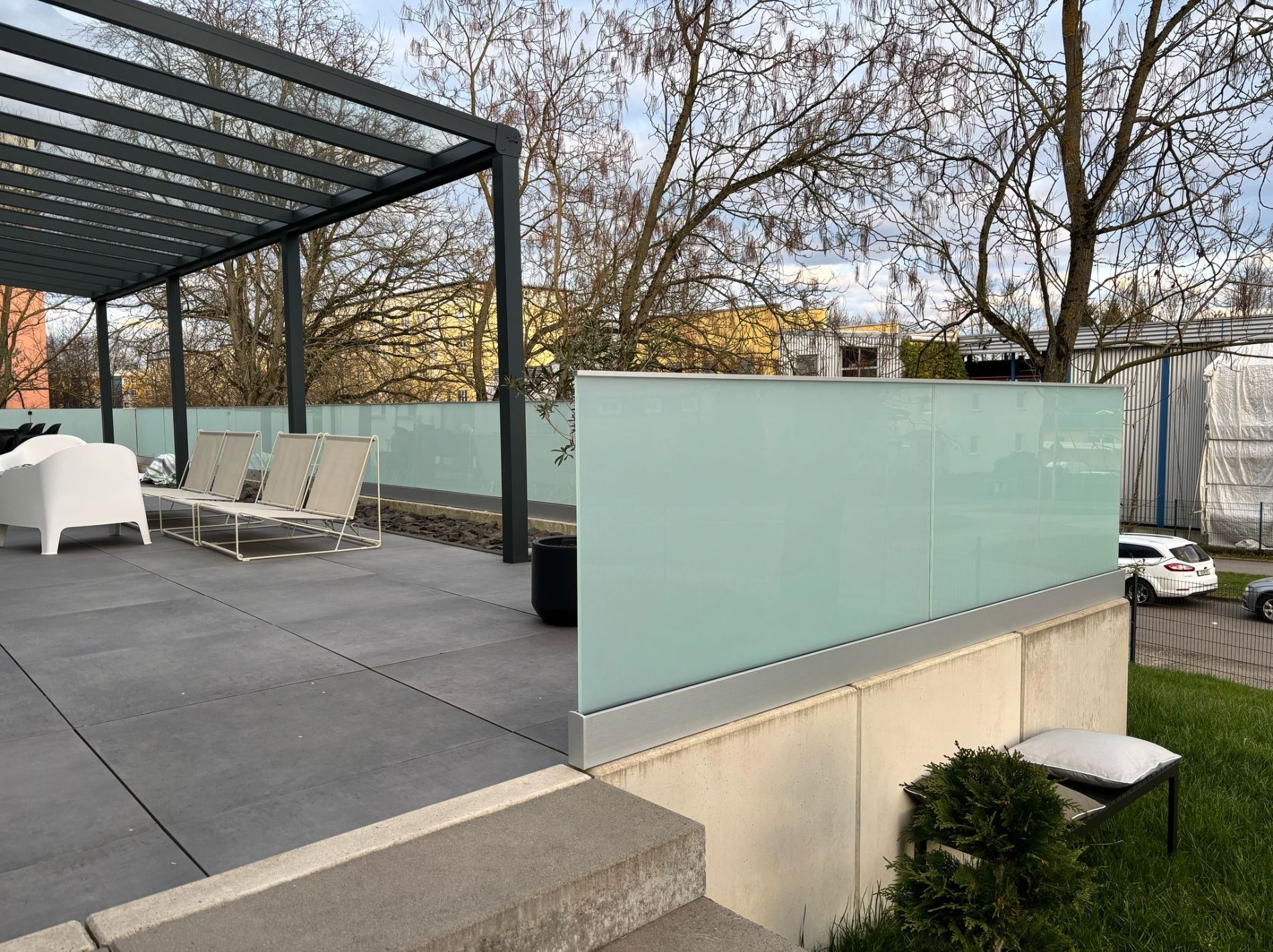 Szklana balustrada z mlecznego szkła, mocowana w aluminiowym profilu, otaczająca taras z szarych płyt, na którym znajdują się białe i beżowe meble ogrodowe; w tle drzewa bez liści i budynki.