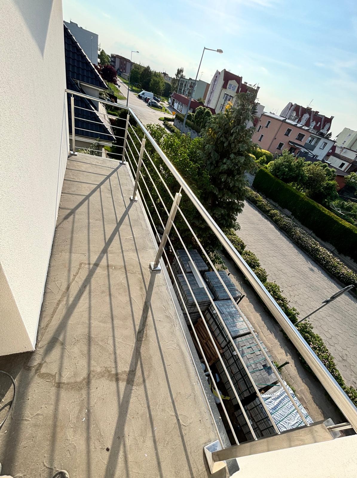 Błyszcząca, stalowa balustrada balkonowa z widokiem na osiedle w słoneczny dzień. Minimalistyczny design, proste linie, nowoczesne wykończenie balkonu.